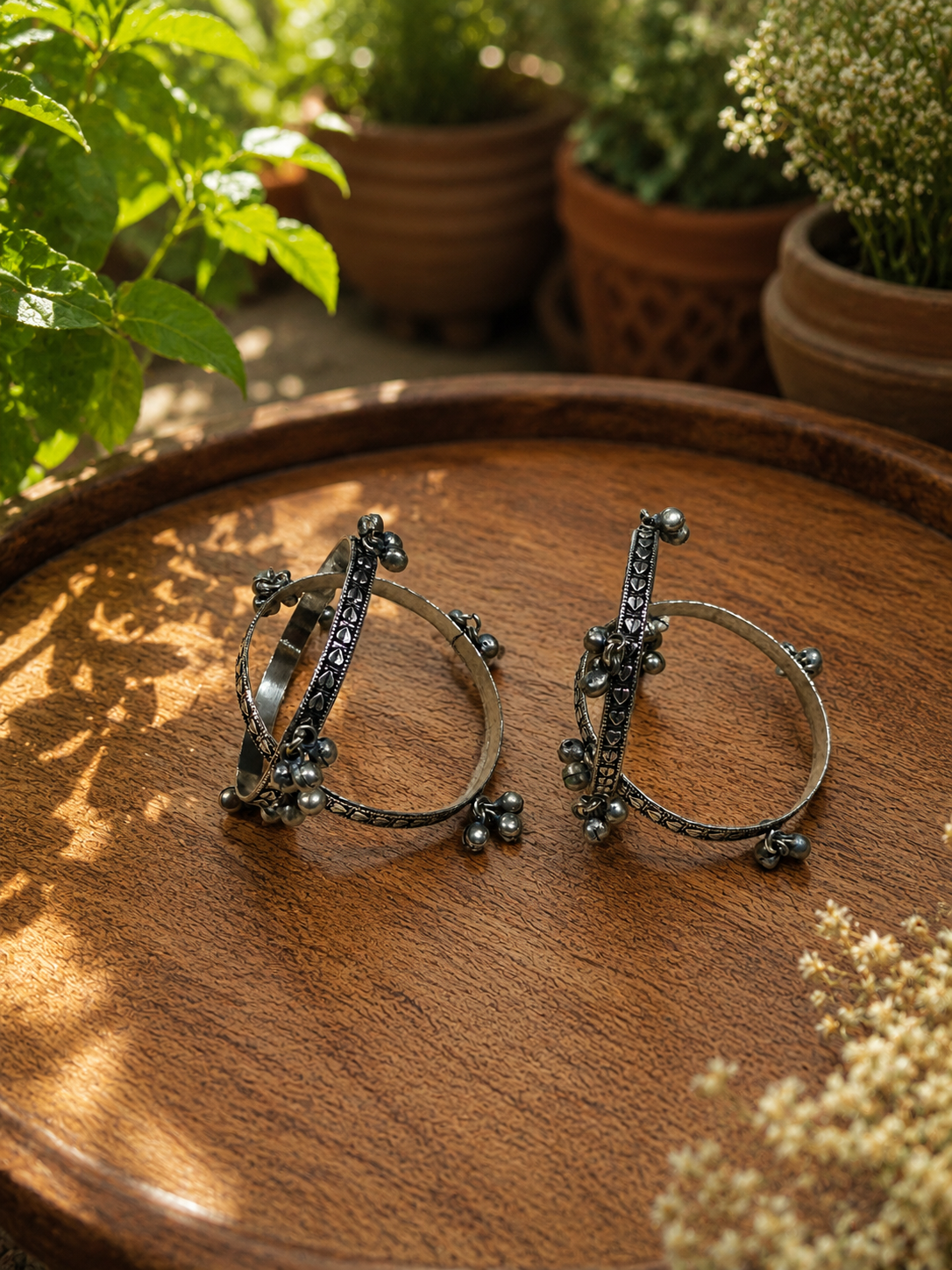 A pair of silver-toned huggie rings with dark oxidized details, displayed on a round wooden tray with a blurred background of potted plants.