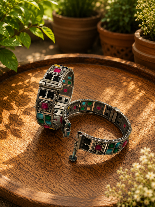 A pair of ornate silver-toned bangles with colorful geometric inlay patterns, displayed on a rustic wooden tray with potted plants in the background.