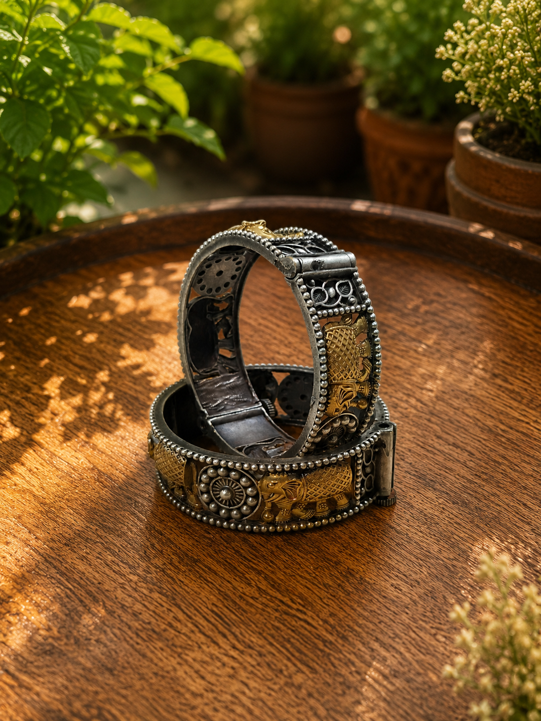 Two ornate silver-toned metal bracelets with gold-toned engraved details and beaded borders, stacked on a round wooden tray with a blurred garden background.