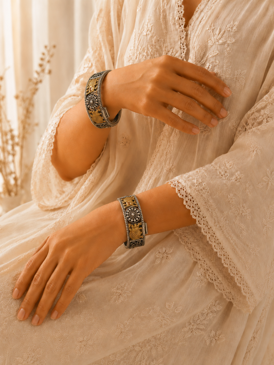 A close-up of a person wearing a cream-colored embroidered dress with lace sleeves, accessorized with two wide metallic bracelets featuring gold and silver patterns.