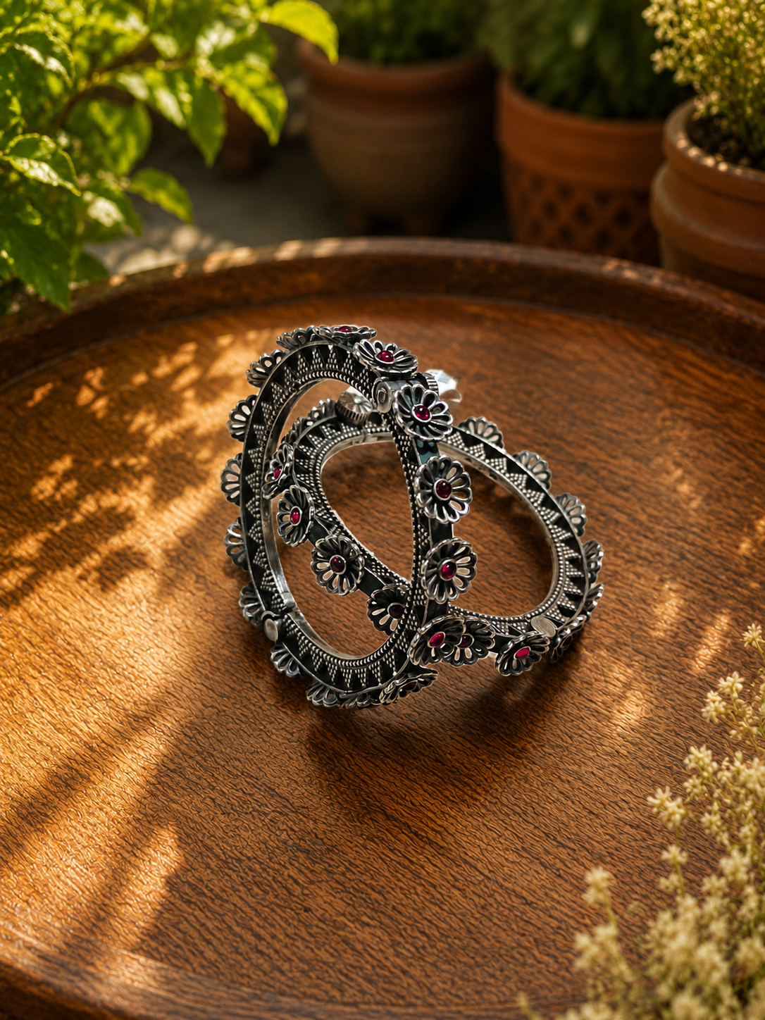 An ornate silver-toned ring with intricate filigree detailing and small red gemstone accents, displayed on a rustic wooden tray with potted plants in the background.