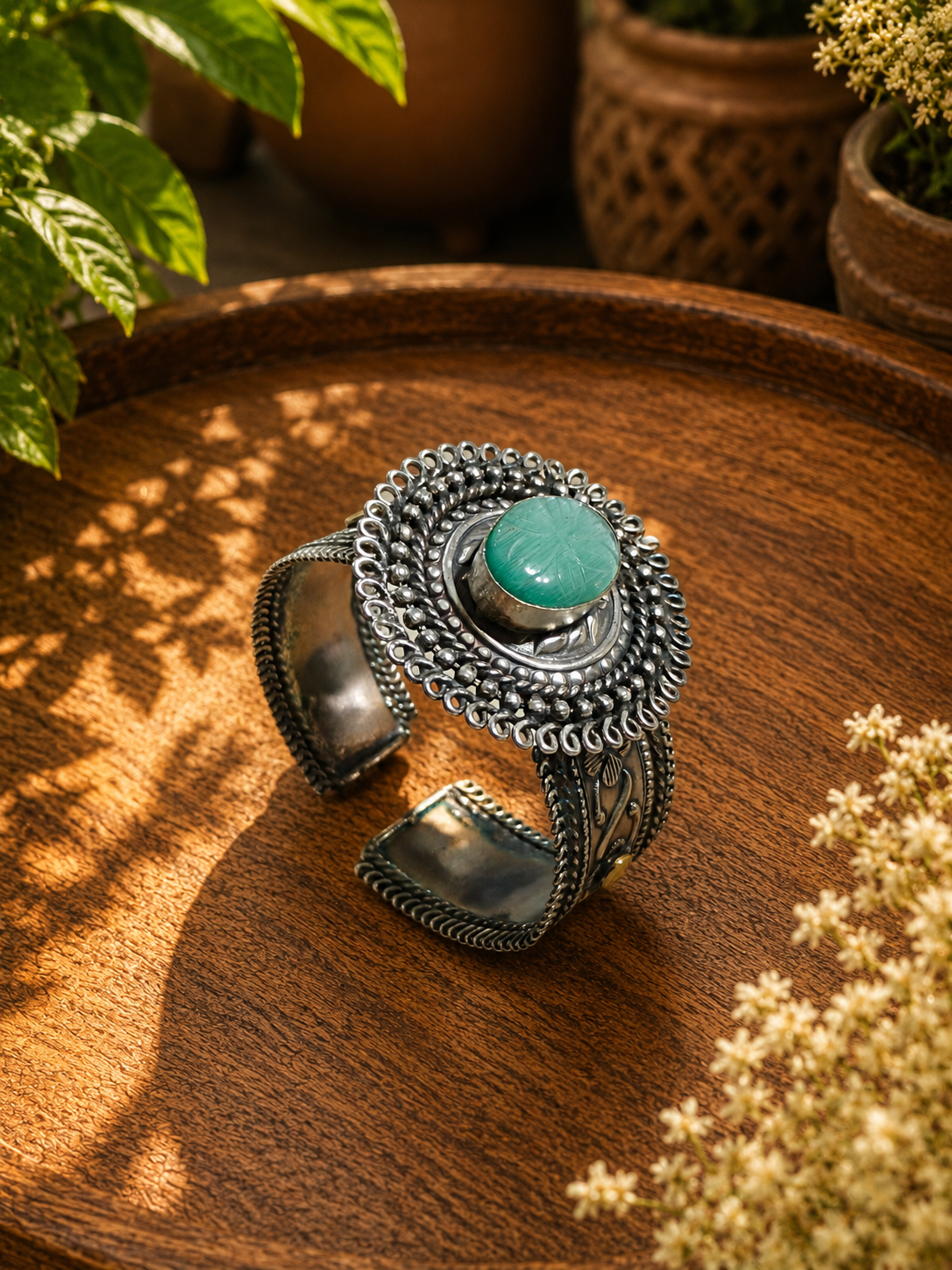 A wide silver cuff bracelet with intricate filigree detailing and a central round green stone, displayed on a wooden tray with plants in the background.