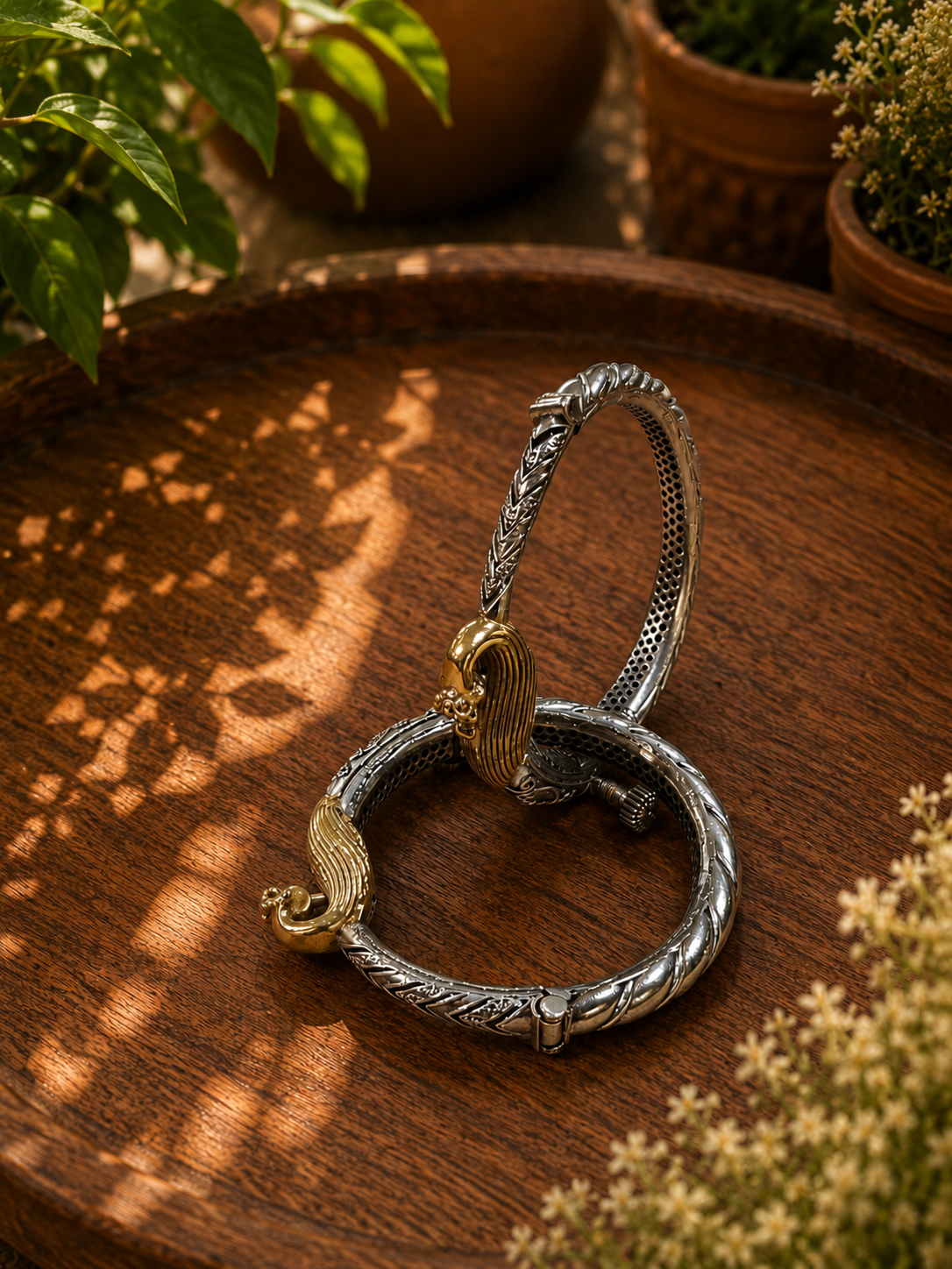 A silver-toned braided bracelet with a gold-toned bird-shaped clasp, displayed on a round wooden tray with warm sunlight and shadow patterns.