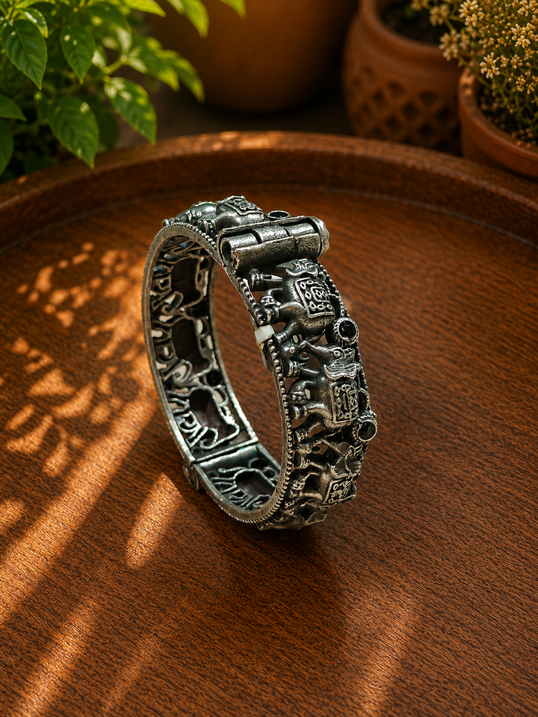 An ornate silver-toned bangle bracelet with detailed relief carvings, displayed on a warm brown wooden surface with soft shadow overlays and blurred potted plants in the background.