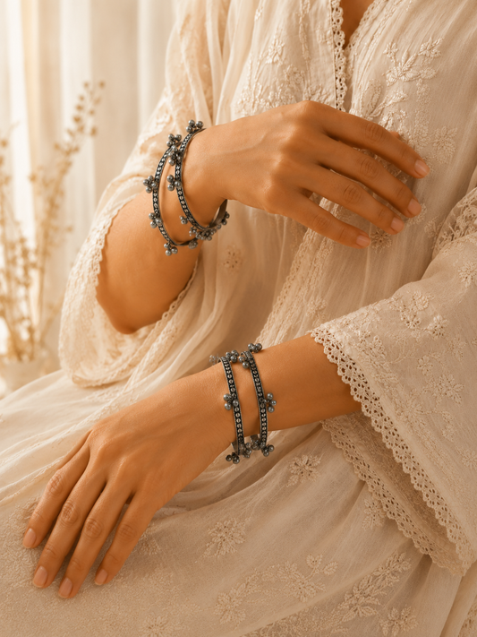 A close-up of a person wearing multiple dark metallic beaded bracelets on both wrists, styled with a cream lace-trimmed dress in a warm, softly lit indoor setting.