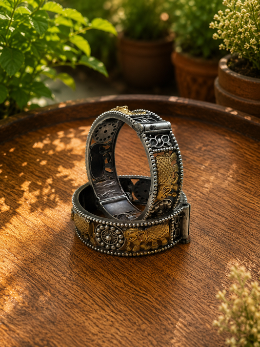 Two ornate silver-toned metal bracelets with gold-toned engraved details and beaded borders, stacked on a round wooden tray with a blurred garden background.