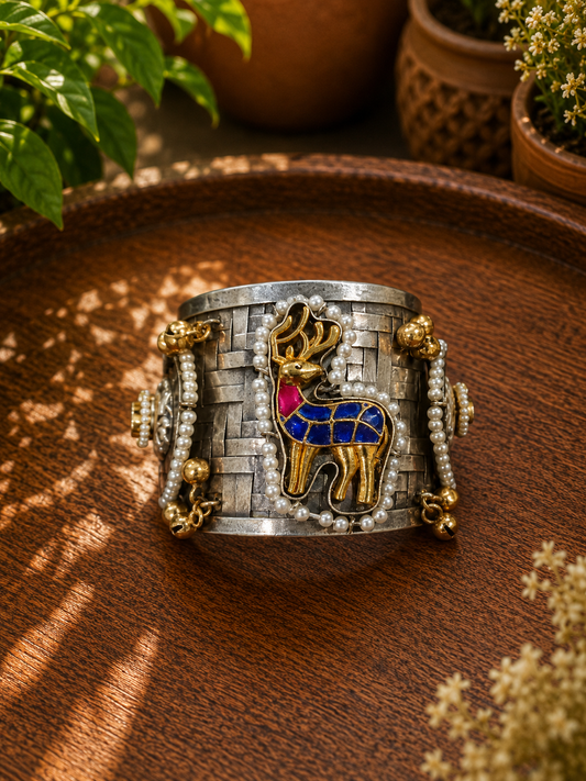 A wide silver-toned cuff bracelet with gold-toned detailing, bead strands, and a central deer motif in gold, blue, and pink, displayed on a wooden surface with plants in the background.