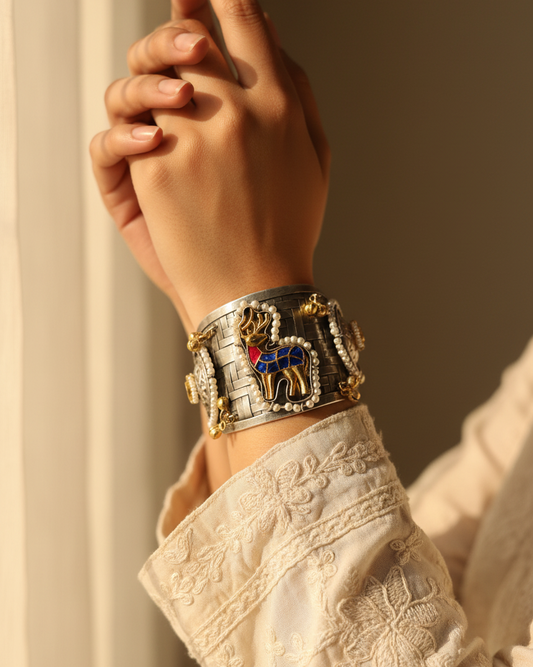 A close-up of a person's wrist wearing a wide silver-toned cuff bracelet adorned with gold-toned details, a central deer motif with blue and red enamel, and small clear stones, paired with a cream embroidered sleeve.