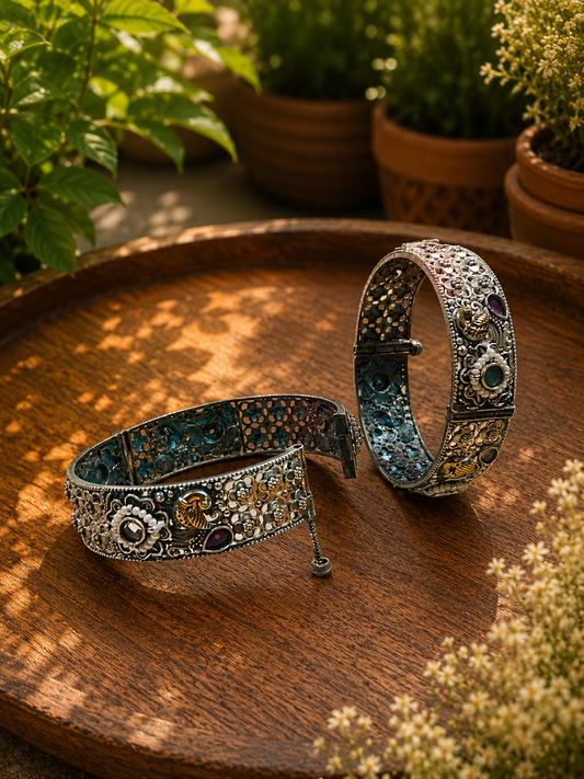 Two ornate silver-toned bangle bracelets with intricate floral engravings and colorful gemstone accents, displayed on a round wooden tray with potted plants in the background.
