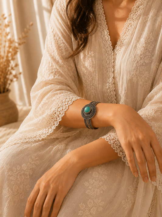 A close-up of a woman wearing a bohemian-style silver-toned bracelet with a round turquoise stone, paired with a cream lace-trimmed maxi dress in a warm, sunlit setting.