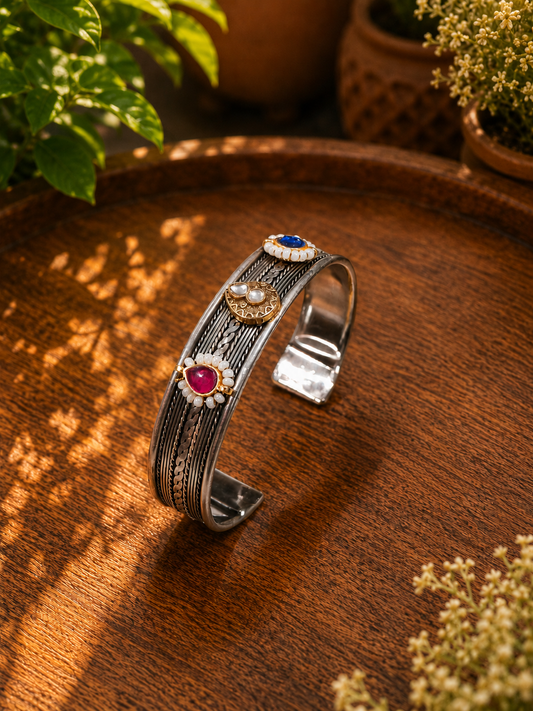 A textured silver cuff bracelet with three decorative gemstone settings, displayed on a rustic wooden surface with soft-focus plants in the background.