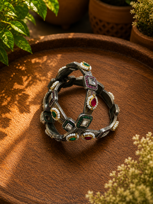A set of dark metallic bangles with colorful gemstone accents, displayed on a rustic wooden tray with green plants in the background.