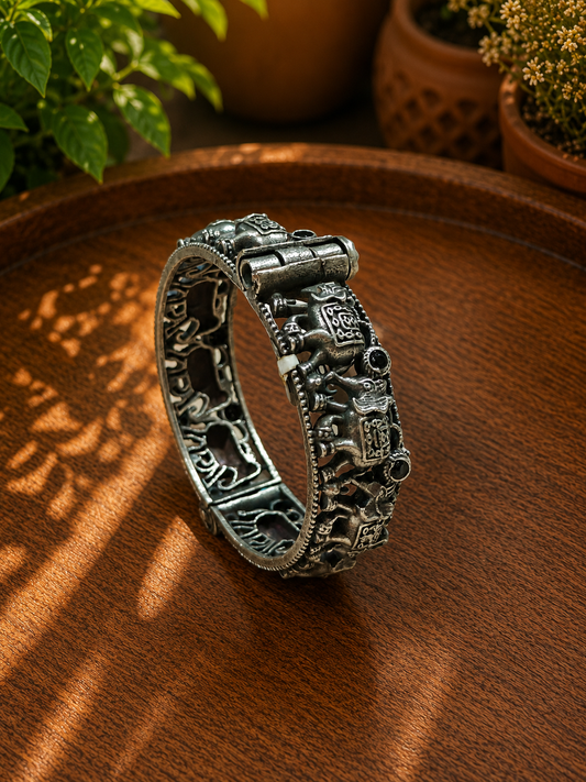 An ornate silver-toned bangle bracelet with detailed relief carvings, displayed on a warm brown wooden surface with soft shadow overlays and blurred potted plants in the background.