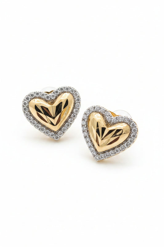 Close-up of premium two-tone heart stud earrings featuring a textured diamond-cut gold center and a silver cubic zirconia halo on black fabric.
